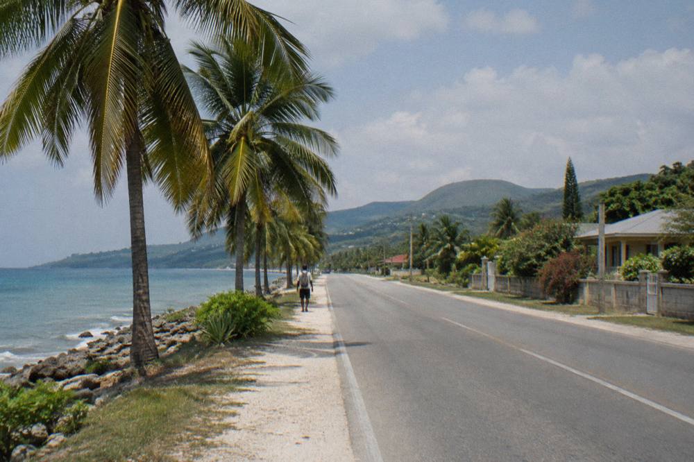 A quiet two-lane coastal highway with palm trees and open sky running toward distant hills