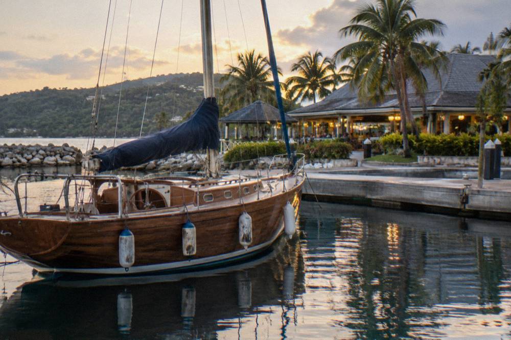 A wooden sailing yacht anchored on still water near a pier with restaurant lights in the background