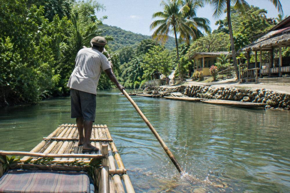 A raft captain poling a bamboo raft along a narrow river between dense green trees