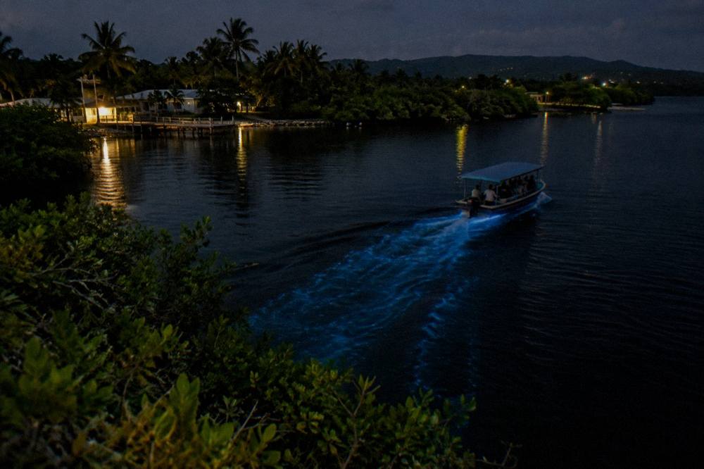 A wooden tour boat moving across dark calm water at night with a faint glow in its wake