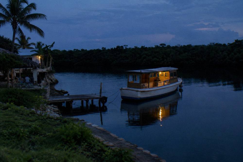 A wooden tour boat resting at a dock on a dark lagoon with mangrove shoreline in the background