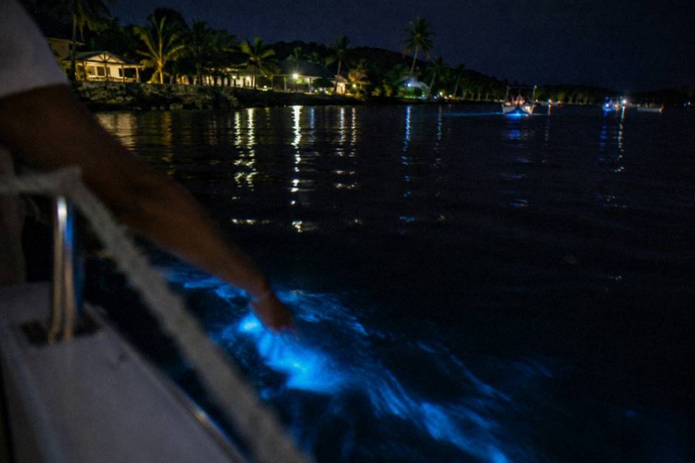 A hand trailing pale blue light through dark water beside a boat rail