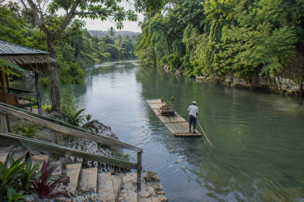 A bamboo raft poled along a wide calm river bordered by tall trees