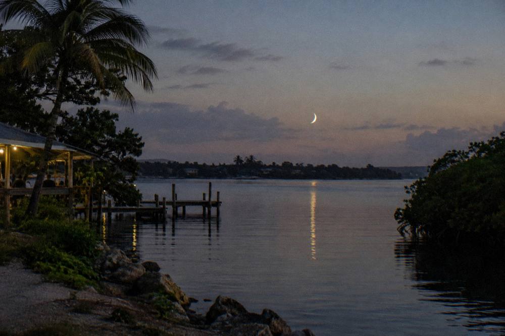 A calm night sky over dark water with a thin crescent moon low above the horizon