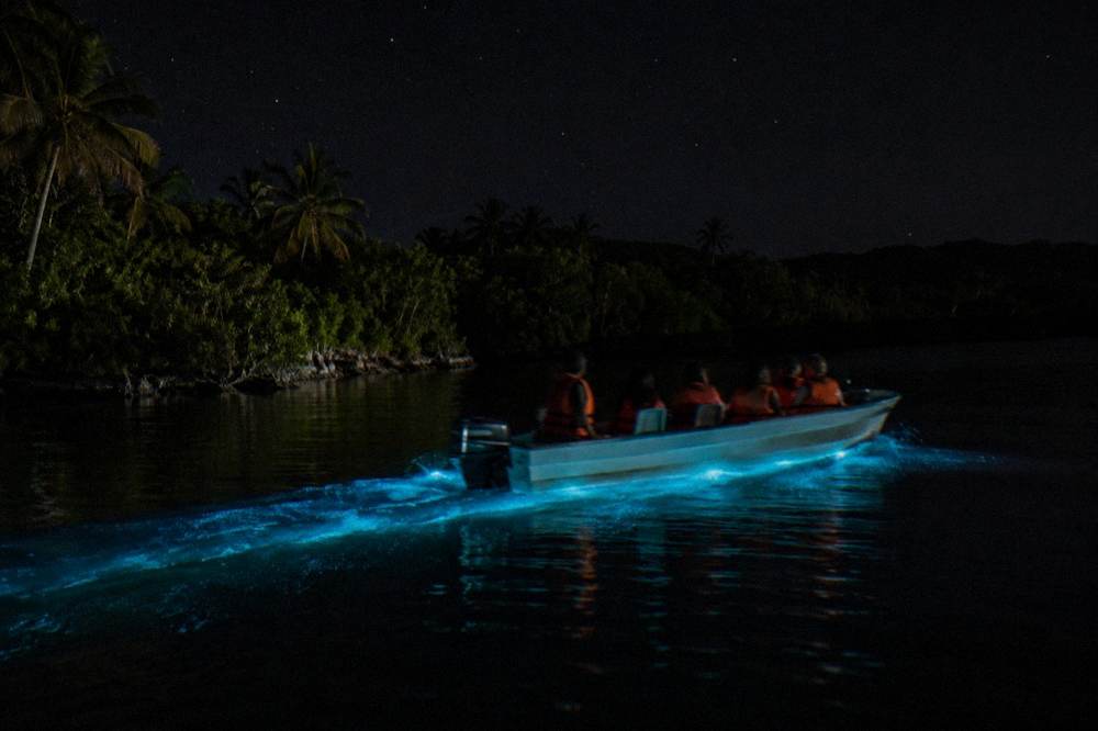 A tour boat with passengers in life jackets crossing dark water with a soft glow trailing behind the hull
