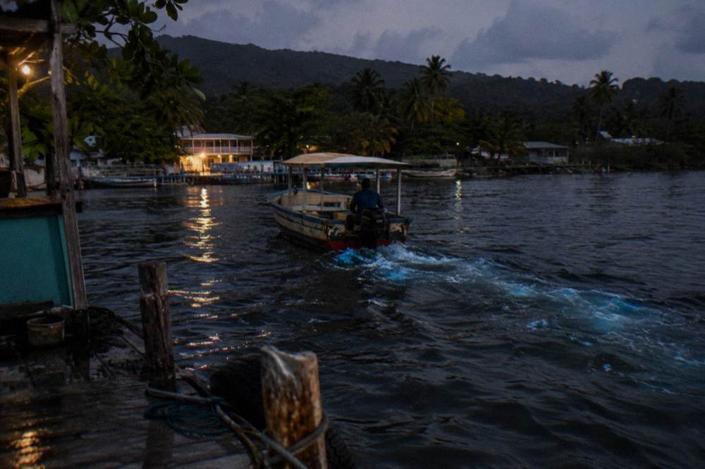 A wooden tour boat moving across dark water leaving a pale blue glowing wake