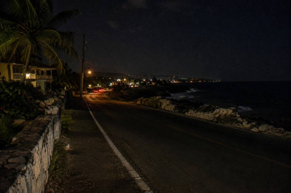 A coastal highway curving along dark shoreline toward distant town lights at night