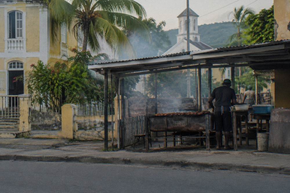 Simple wooden table at an open-air jerk chicken restaurant with smoke drifting from a grill